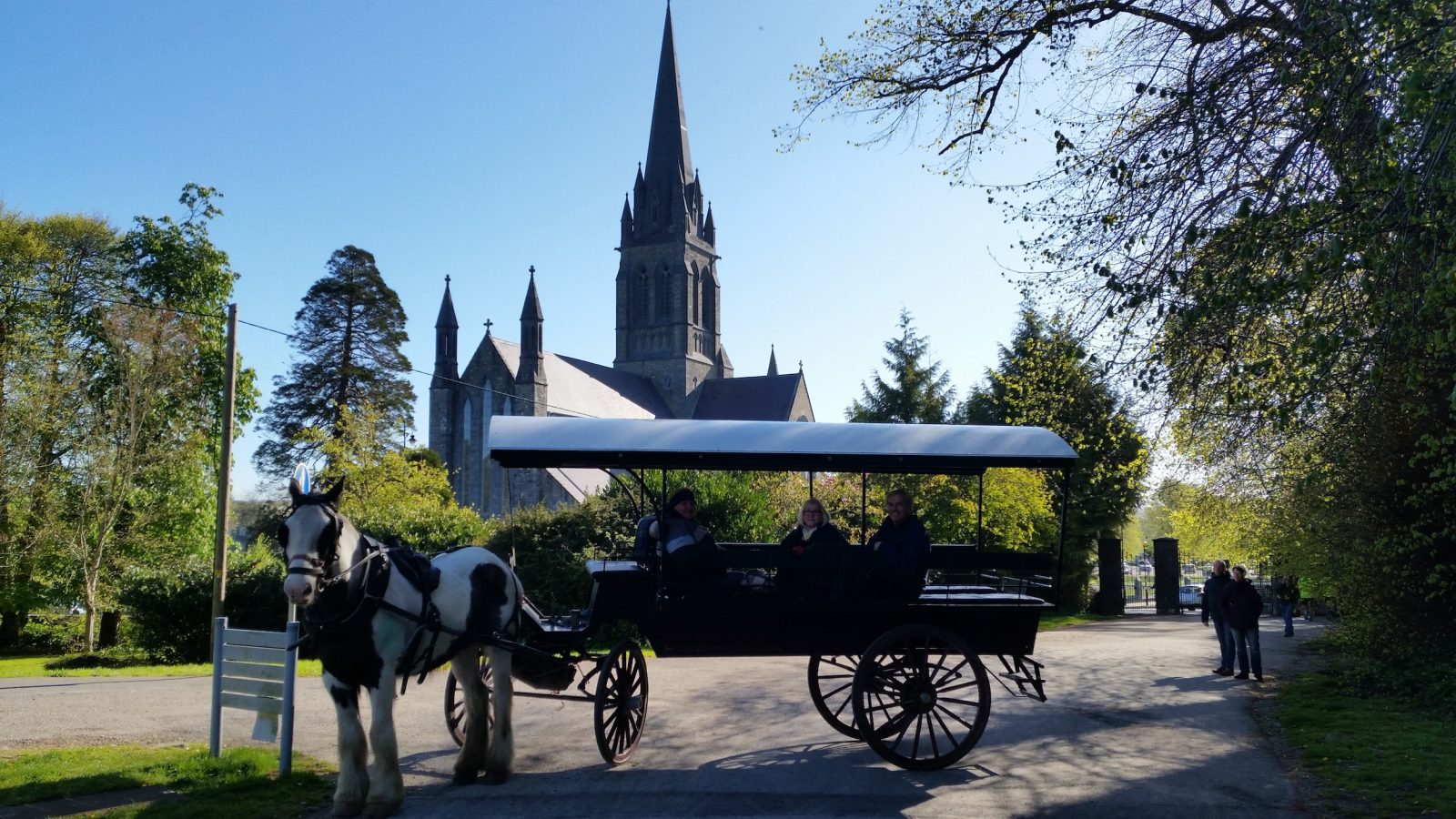 A horse-drawn carriage with people inside is parked on a tree-lined path. In the background, a large church with a tall spire is visible under a clear blue sky. Passersby and lush greenery surround the area.