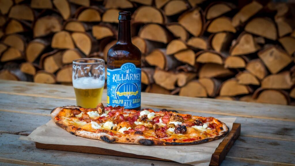 A pizza topped with various ingredients is placed on a wooden board. Beside it is a half-filled glass and a bottle of beer with a blue label. In the background, neatly stacked firewood is visible.