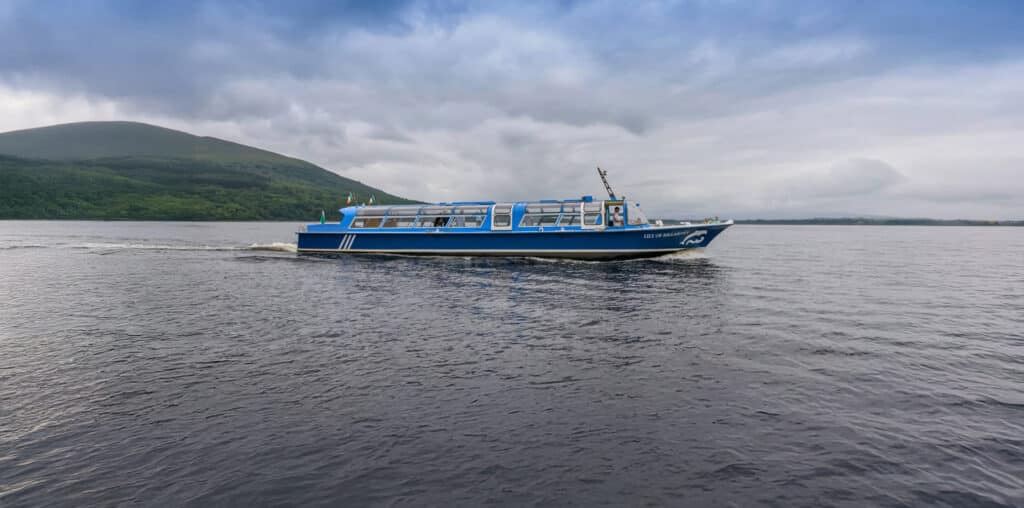 A blue tour boat sails on a lake under a cloudy sky, with a forested hill in the background.