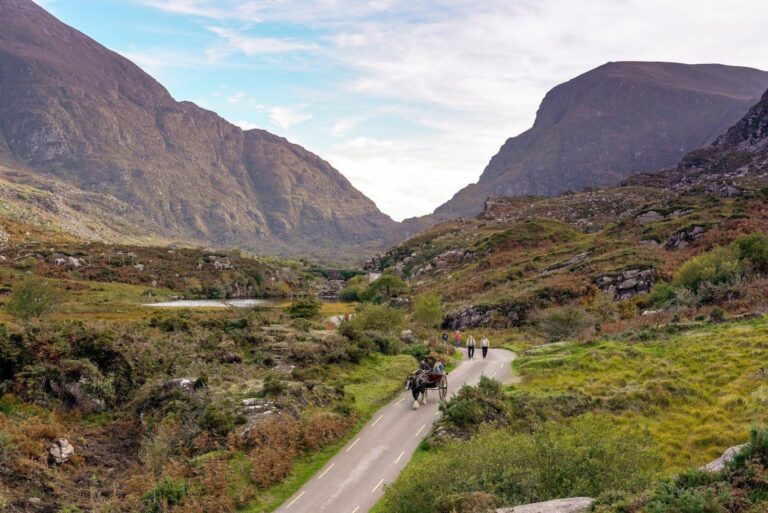 A scenic valley with towering mountains in the background. A narrow road winds through lush greenery, and a horse-drawn carriage is accompanied by two people walking beside it. The sky is partly cloudy, adding to the picturesque landscape.