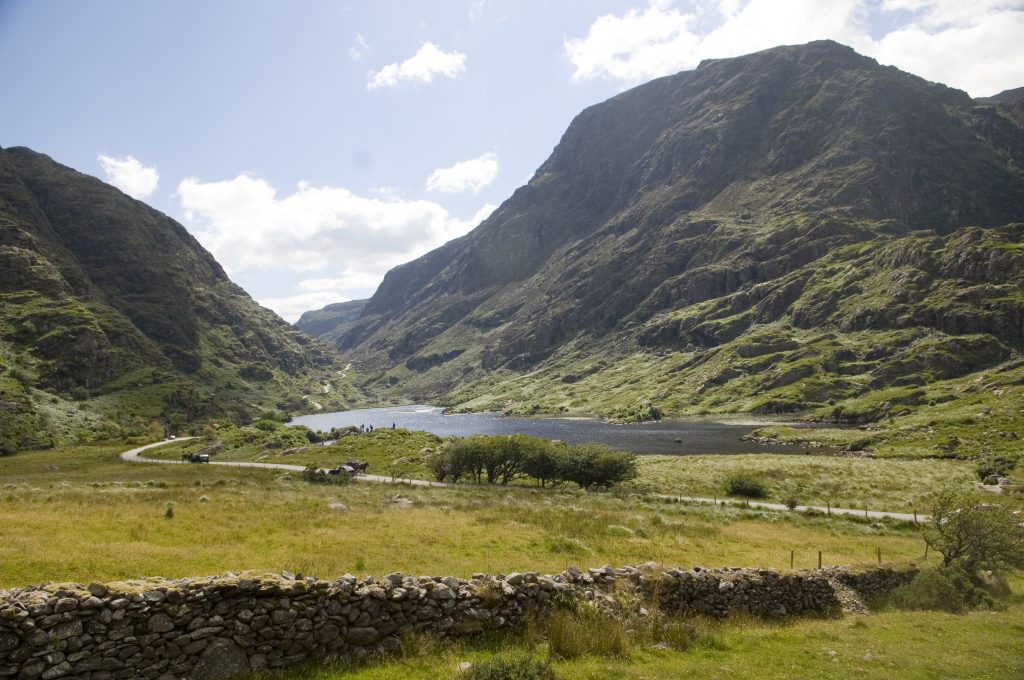 Scenic view of a tranquil valley with a lake surrounded by lush green fields and steep, rocky hills under a partly cloudy sky. A stone wall and small trees dot the landscape, creating a peaceful and natural atmosphere.