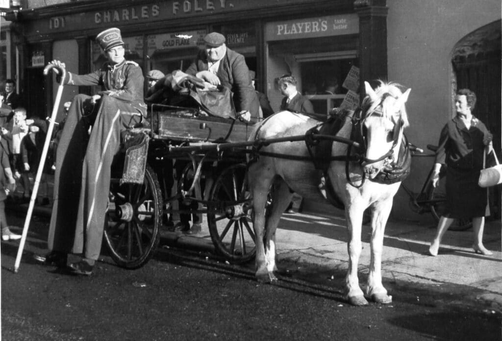 A vintage black and white photo shows a horse-drawn cart on a street. A person in a uniform stands beside the cart, while another person sits at the back. Pedestrians walk by, and old shop signs are visible in the background.