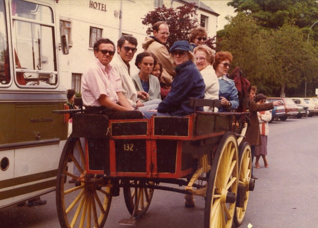 A group of people sit on a horse-drawn carriage with red and yellow wheels on a street lined with parked cars. A hotel building is visible in the background. The passengers appear to be tourists enjoying a ride.