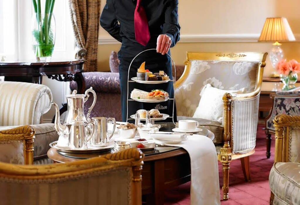 A waiter serves an elegant afternoon tea set on a three-tiered stand in a luxurious lounge. The setting includes plush armchairs, a wooden table with a white runner, a silver tea set, and a lamp in the background.