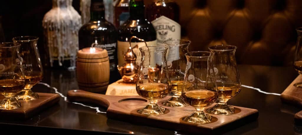 A selection of whiskey glasses filled with amber liquid sits on a wooden flight board. Several whiskey bottles and a small barrel are in the background on a dark, marble-topped table. The setting is warmly lit, creating a cozy atmosphere.