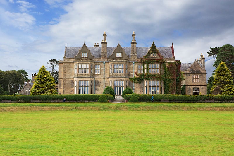 Grand stone manor with ivy, surrounded by lush lawn and trees under a cloudy sky.