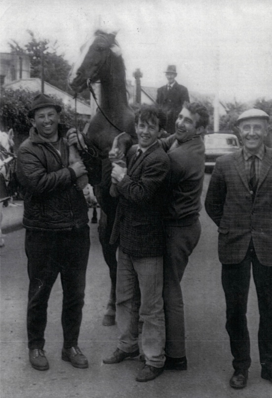 Four men smiling with a horse on a street; another man and horse in the background.