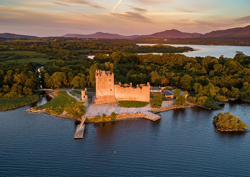 Aerial view of a lakeside castle surrounded by greenery and mountains at sunset.