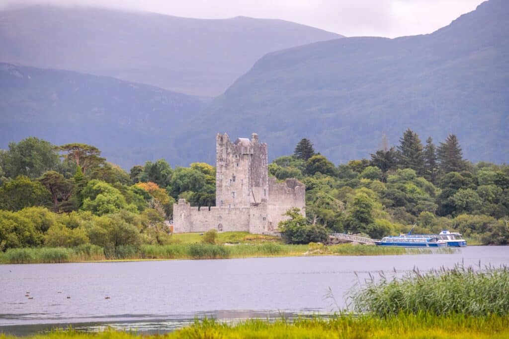 A castle by a lake with mountains in the background and a boat nearby.