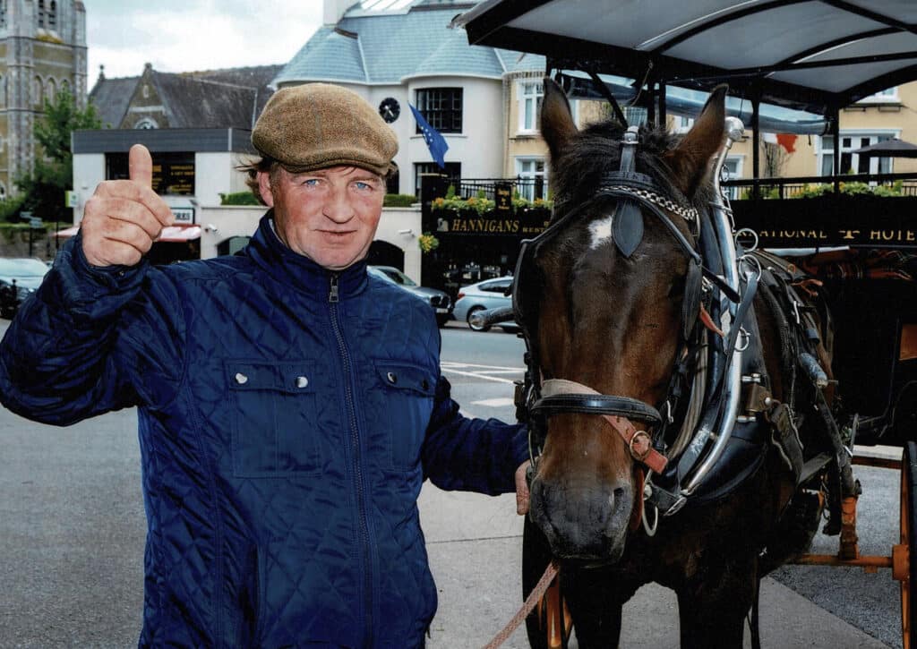 Man in a cap gestures with a horse-drawn carriage in a city street.