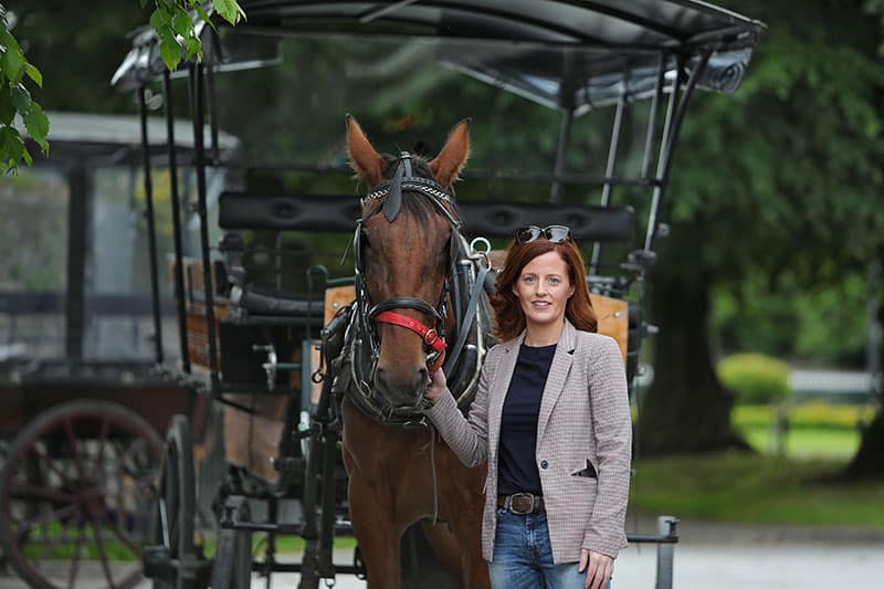 Woman standing with a horse in front of a carriage in a green outdoor setting.