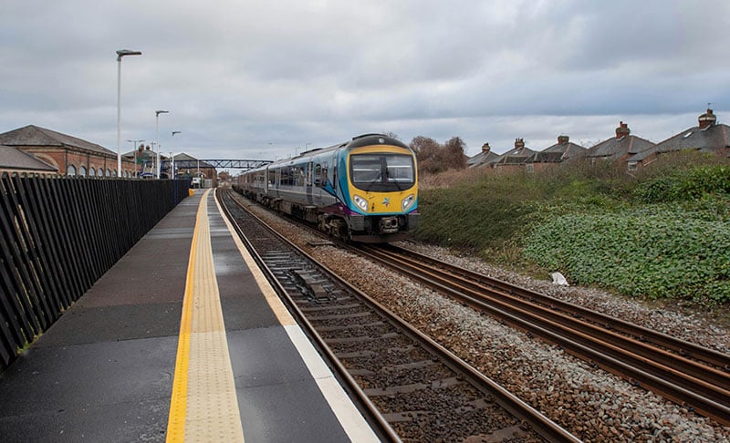 Train approaching a station platform on a cloudy day, with houses in the background.