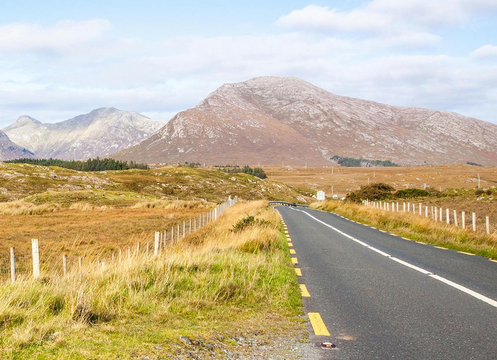 Road leading to mountains under a cloudy sky, with grassy fields on both sides.