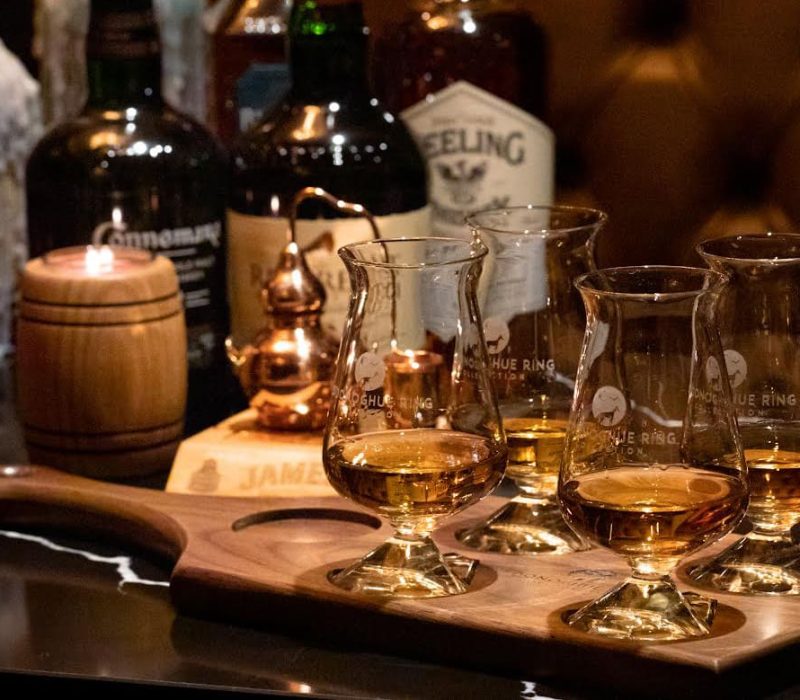 A selection of whiskey glasses filled with amber liquid sits on a wooden flight board. Several whiskey bottles and a small barrel are in the background on a dark, marble-topped table. The setting is warmly lit, creating a cozy atmosphere.