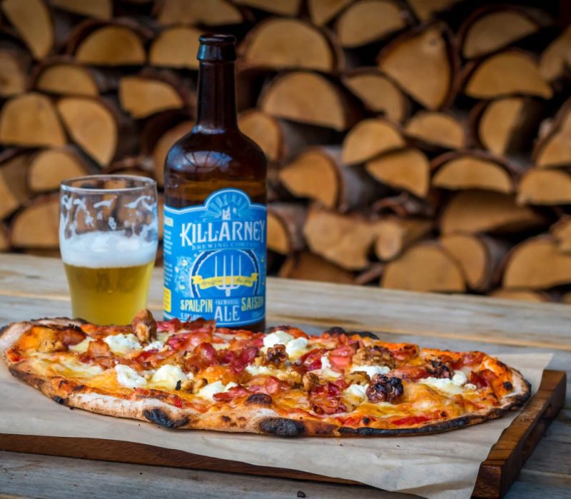 A pizza topped with various ingredients is placed on a wooden board. Beside it is a half-filled glass and a bottle of beer with a blue label. In the background, neatly stacked firewood is visible.