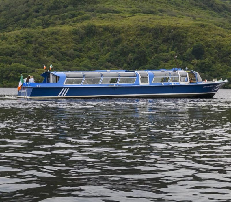 A blue tour boat on a lake surrounded by green hills.