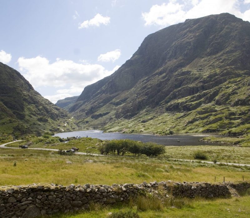 Scenic view of a tranquil valley with a lake surrounded by lush green fields and steep, rocky hills under a partly cloudy sky. A stone wall and small trees dot the landscape, creating a peaceful and natural atmosphere.