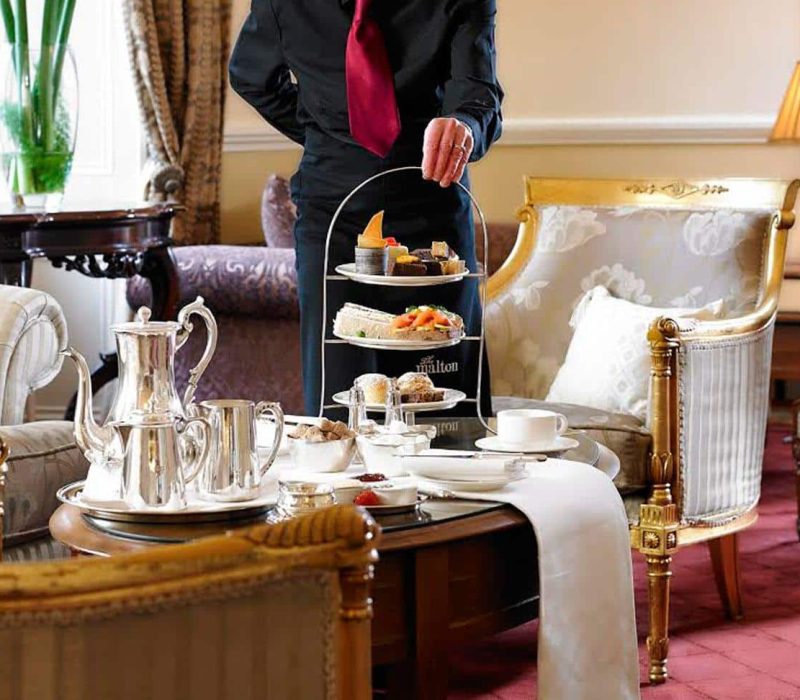 A waiter serves an elegant afternoon tea set on a three-tiered stand in a luxurious lounge. The setting includes plush armchairs, a wooden table with a white runner, a silver tea set, and a lamp in the background.